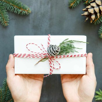 Close up of man hands holding white wrapped gift box with ribbon, sprig of conifer, cone on black background. Winter holiday pattern. Top view. Christmas background.